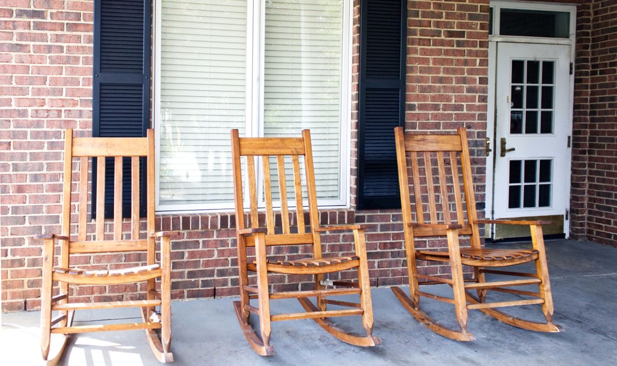 Three wooden rocking chairs sit on a porch in front of a brick wall and windows with shutters.