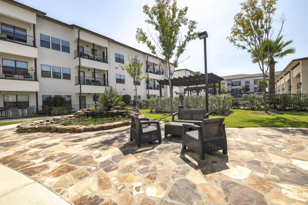 An outdoor patio area with seating and landscaping in front of a multi-story building on a sunny day.