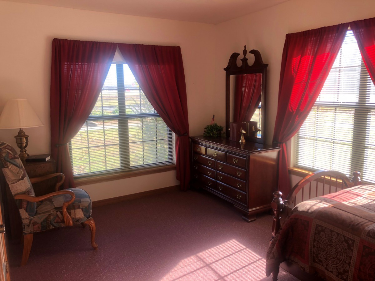 A cozy bedroom featuring a window with red curtains, a wooden dresser with a mirror, and a comfortable chair.