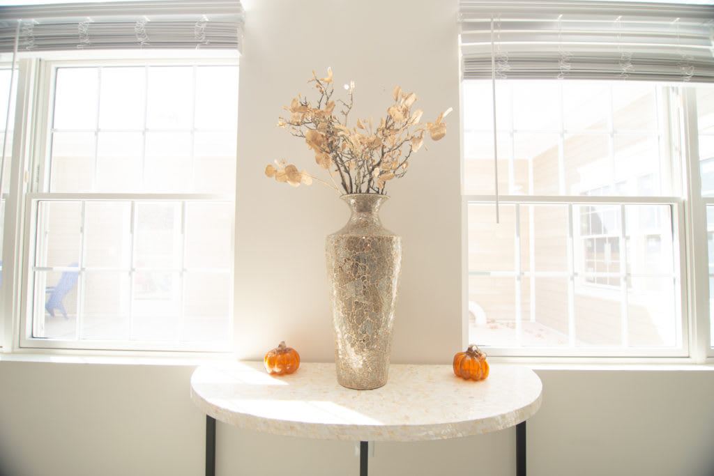 A minimalist white table with a decorative vase of dried flowers and two small pumpkins.