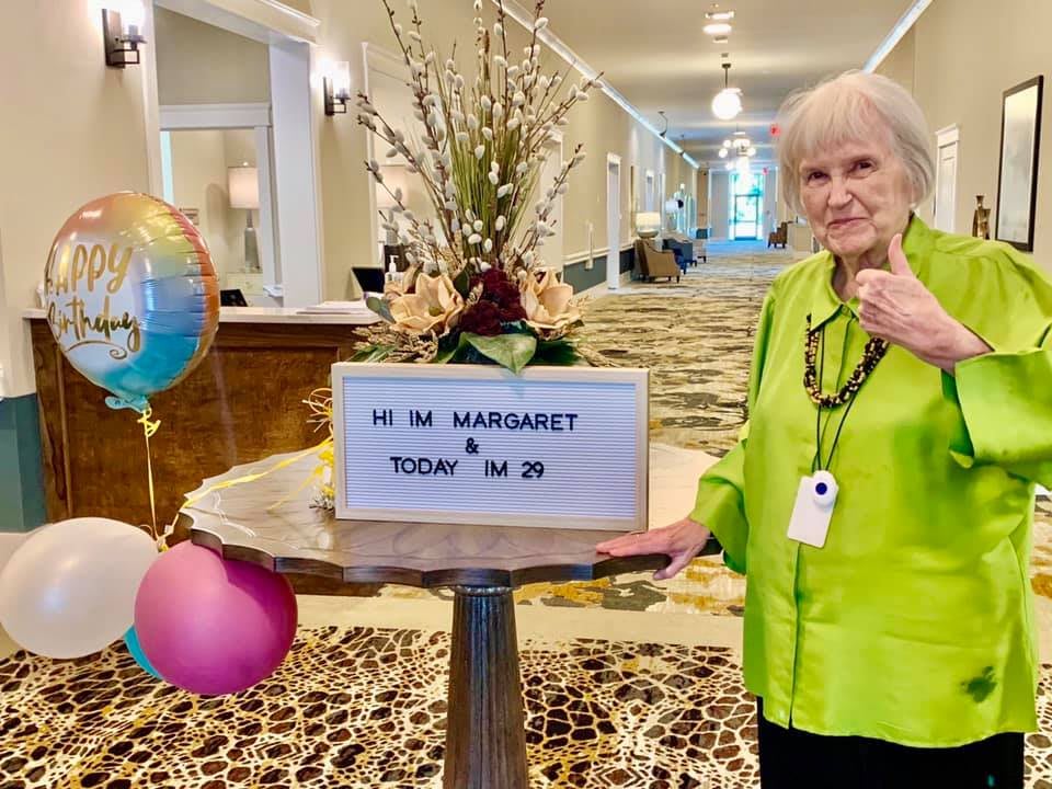 A woman standing next to a sign celebrating her birthday with balloons and flowers in a hallway.