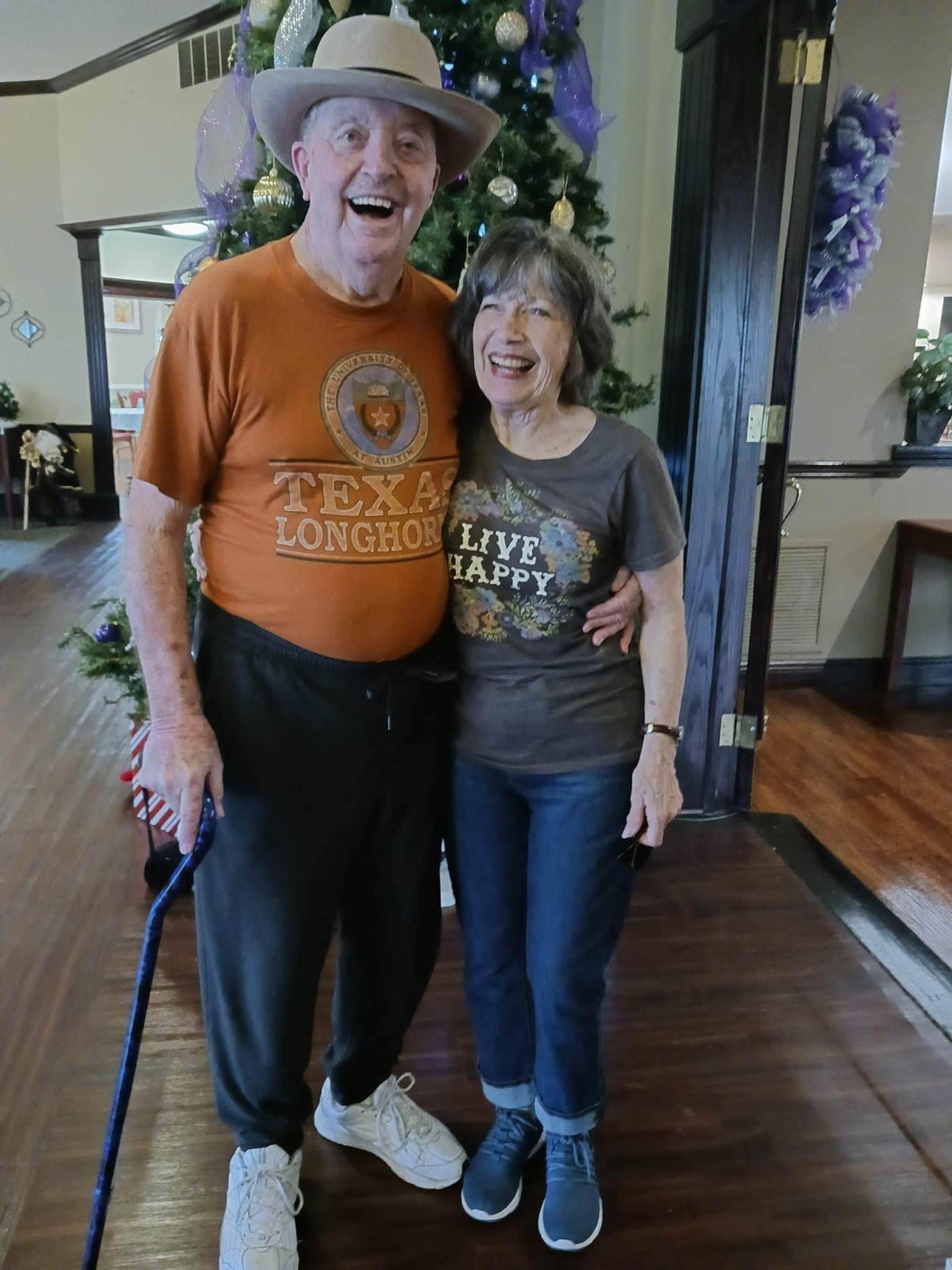An elderly couple smiling and posing together indoors, with a Christmas tree in the background.