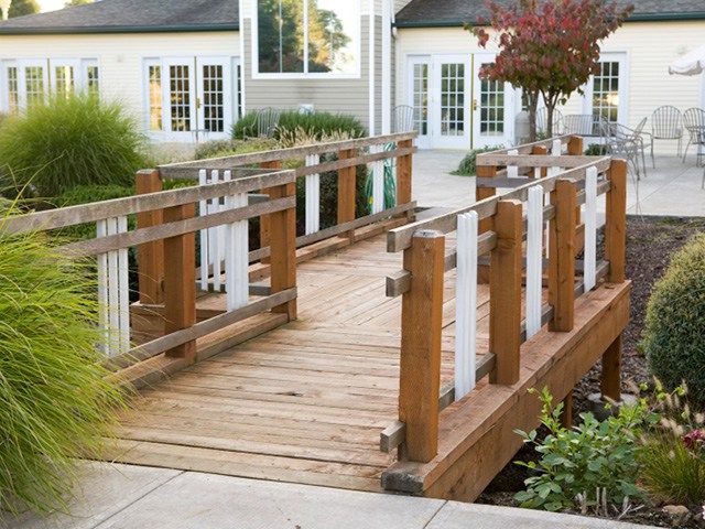 A wooden bridge in a garden setting leads to a building with large windows.
