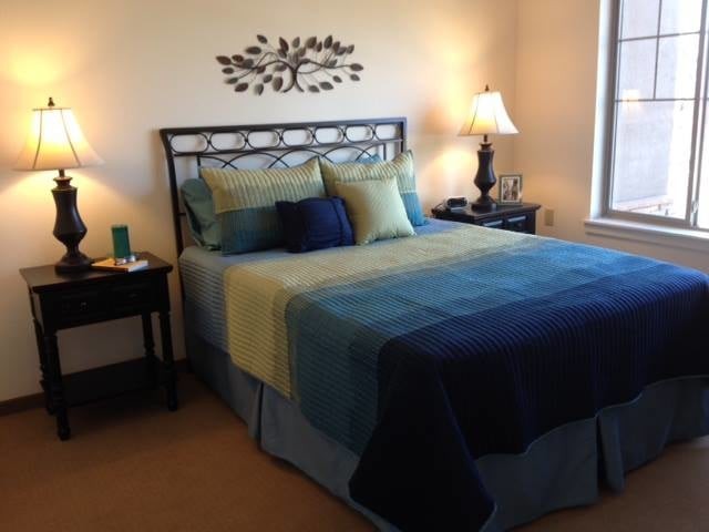A cozy bedroom with a neatly made bed, accented with blue and green pillows, a nightstand with a lamp, and decorative wall art.