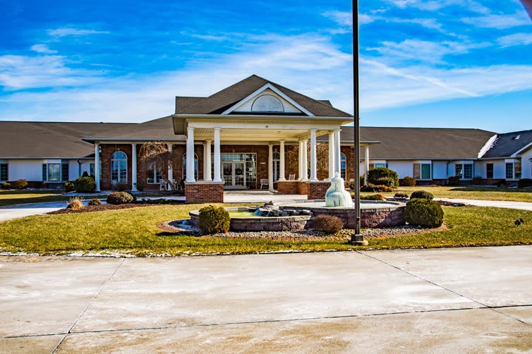 A large building with a grand entrance featuring columns, surrounded by landscaped gardens and a fountain under a clear blue sky.