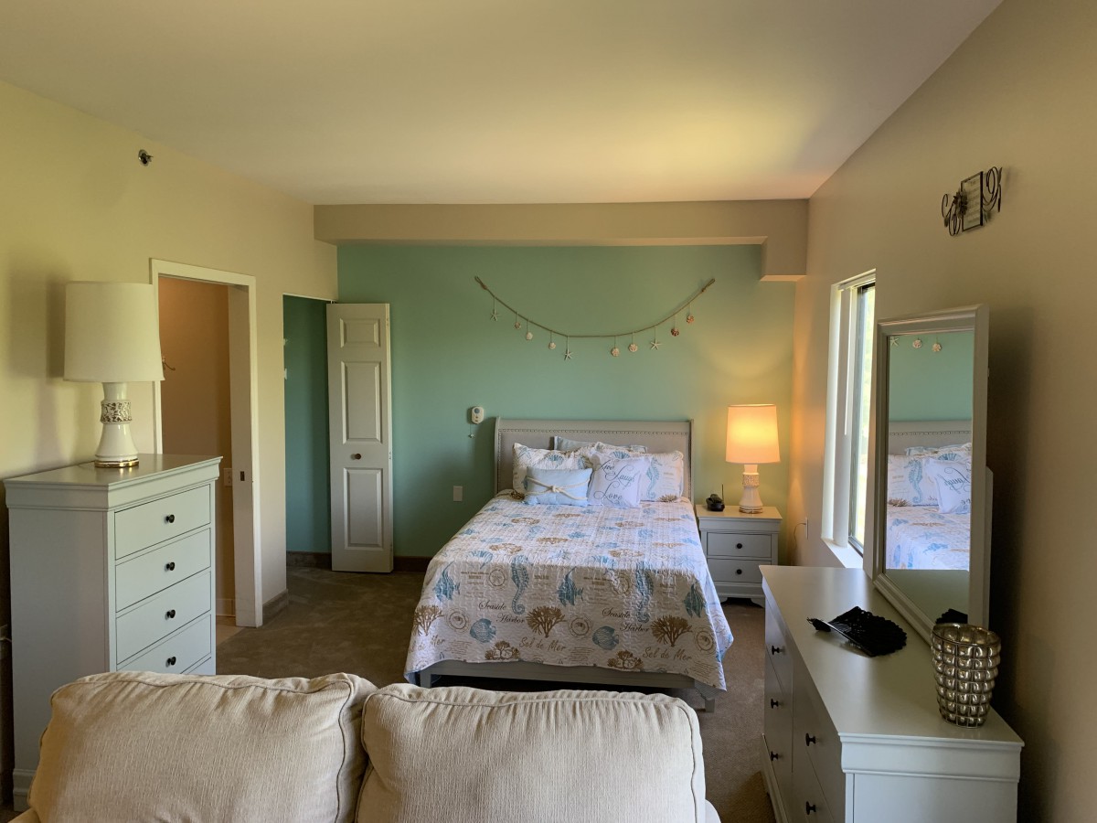 A cozy bedroom with pastel colors, featuring a bed, dresser, and mirror.
