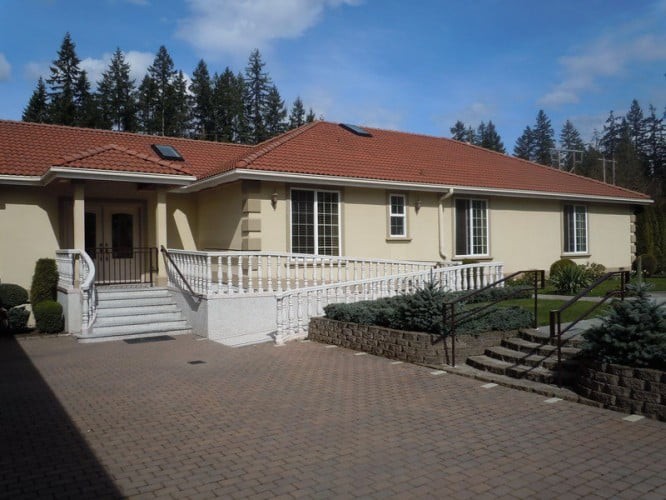A single-story residential house with a red roof, featuring a paved driveway and surrounded by trees.