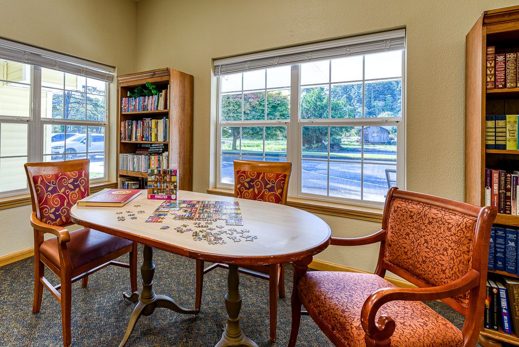 A cozy room with a puzzle on the table, surrounded by chairs and a bookshelf by the window.