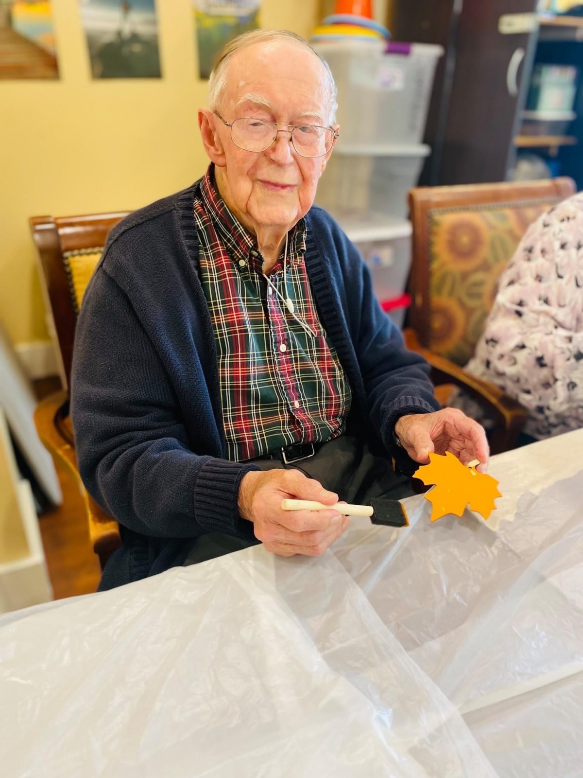 An elderly man sits at a table, holding an orange autumn leaf while engaging in a crafting activity indoors.