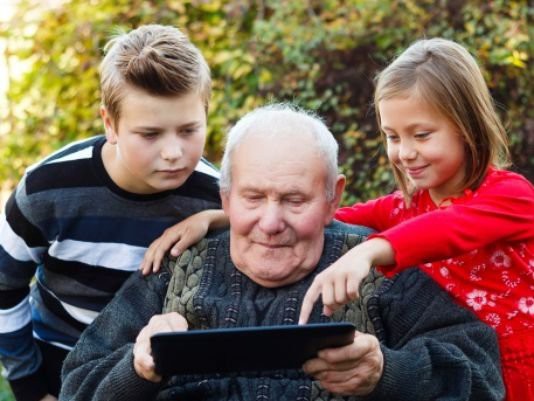 A grandfather shares a moment with his grandchildren, exploring a tablet together outdoors.