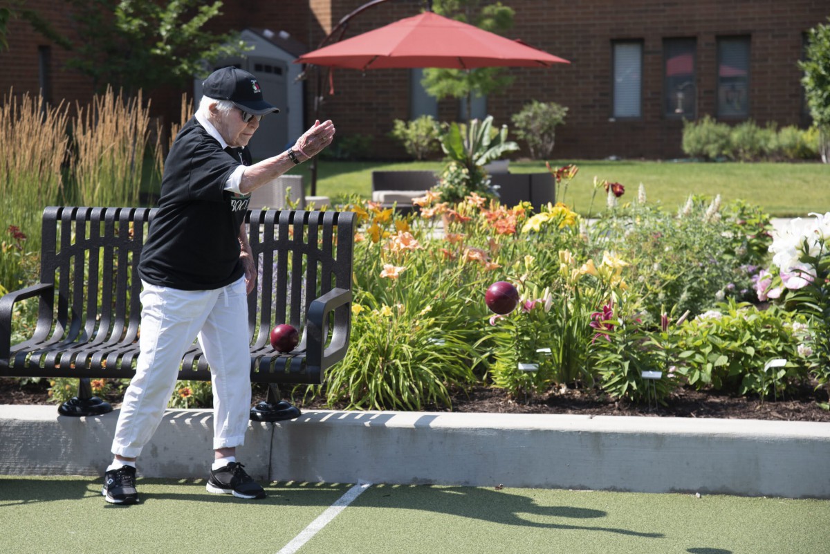 An elderly person playing bowling outdoors near a garden on a sunny day.