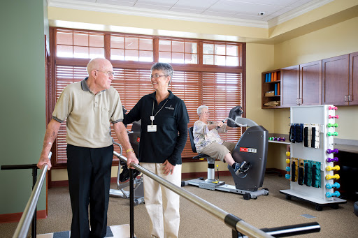 An elderly man receives assistance from a therapist in a rehabilitation center while another senior engages with exercise equipment.
