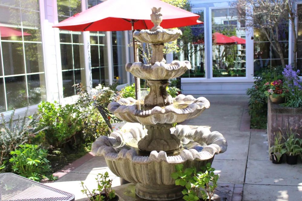 A multi-tiered stone fountain with flowing water situated on a sunlit patio next to a building, surrounded by greenery and under the shade of red umbrellas.