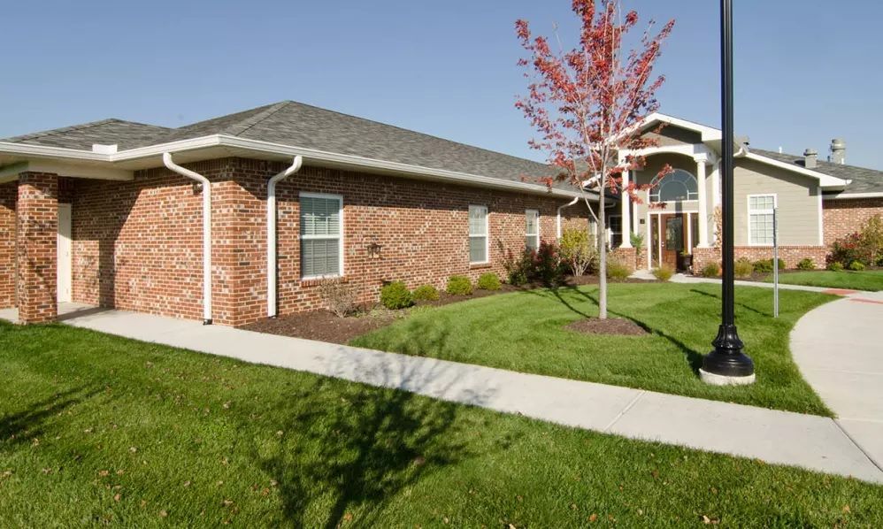 A suburban residential building with brick facade and white columns, surrounded by a landscaped front yard.