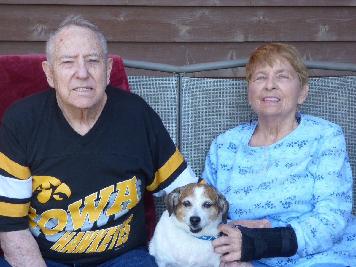 An elderly couple sitting together with their dog on a porch.