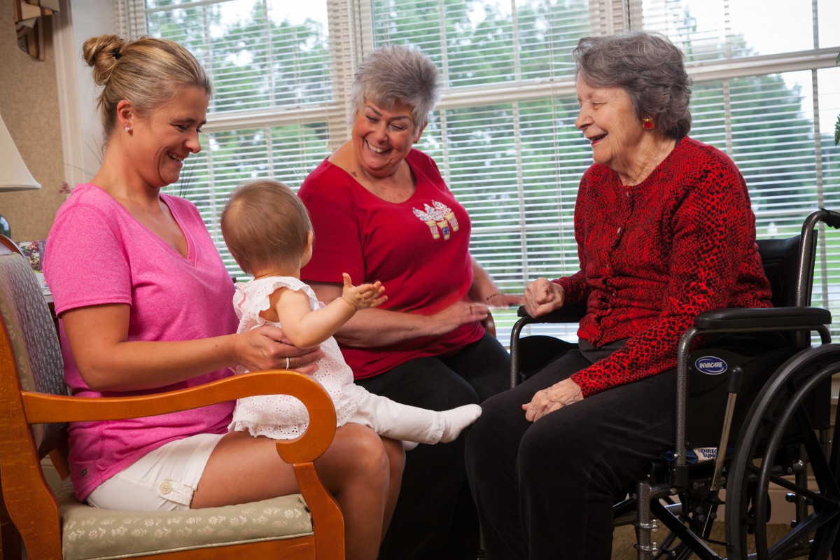 A multi-generational family gathering with a baby interacting with two older women and one younger woman indoors.