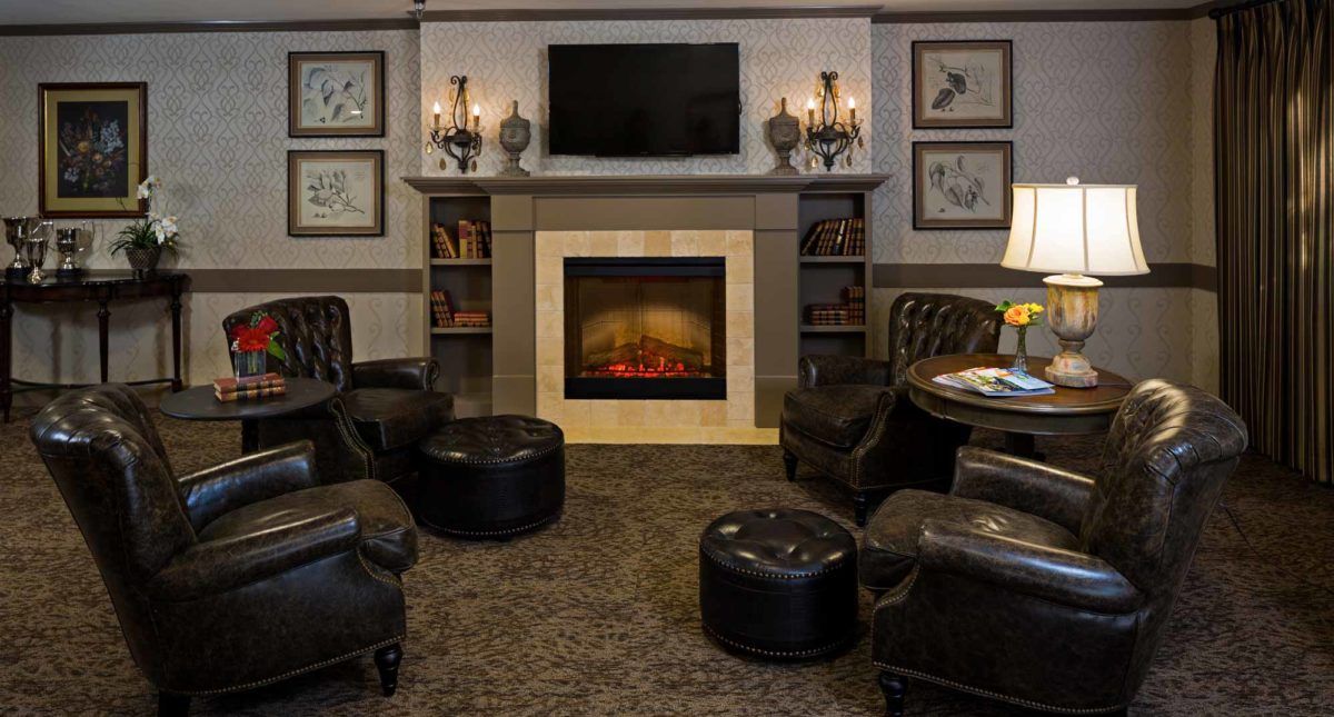 A cozy living room with a fireplace surrounded by leather chairs and ottomans, topped with a television and adorned with decorative sconces and framed art.