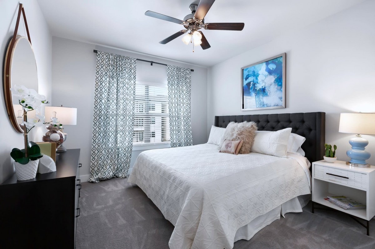 A modern bedroom with a large bed, white bedding, and a ceiling fan. The room features patterned curtains, a piece of abstract artwork above the bed, and a lamp on the nightstand.