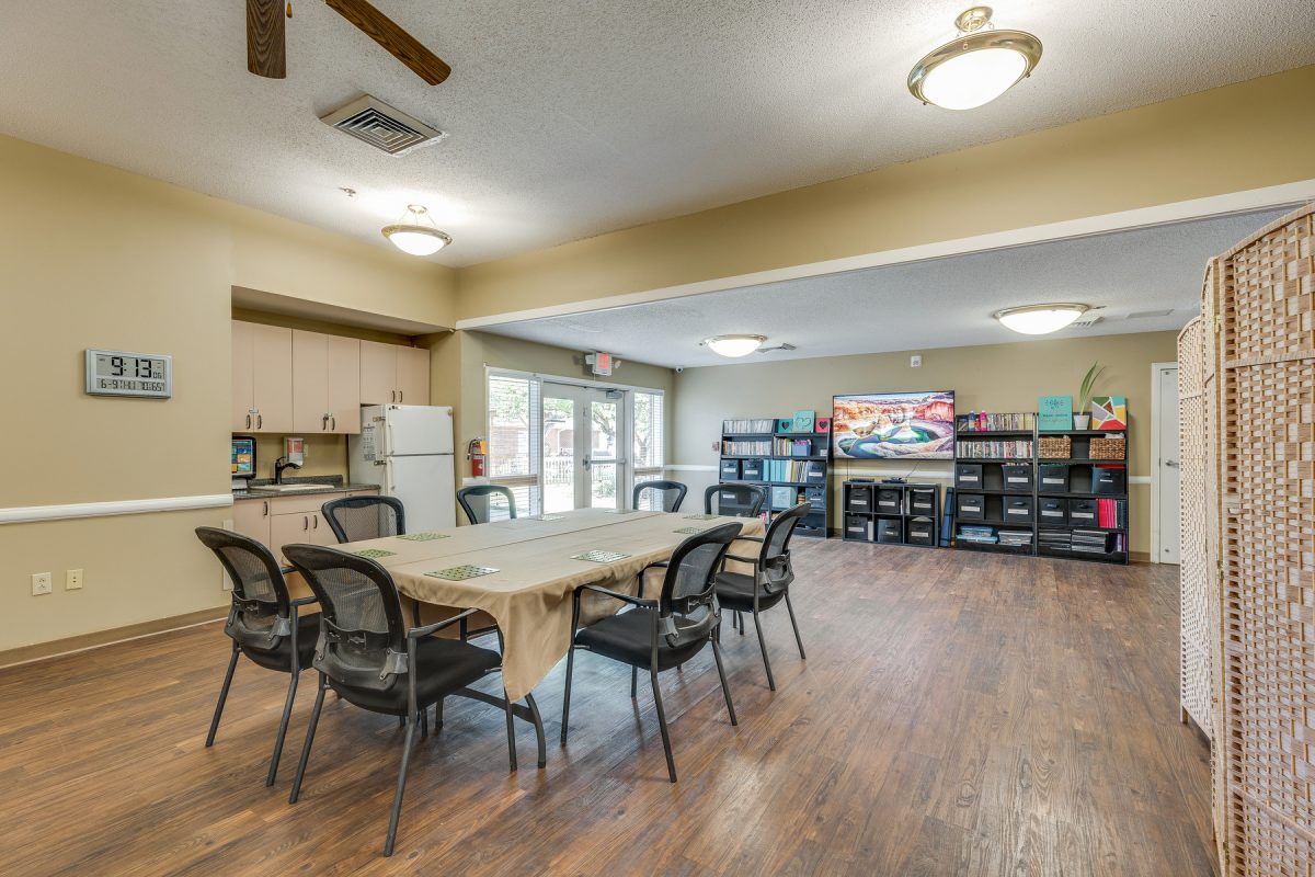 A community room featuring a table and chairs, a kitchen area with a refrigerator, bookshelves, and a television.