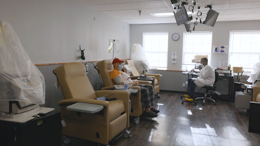 A patient sits in a comfortable chair in a medical treatment area, surrounded by other empty chairs.