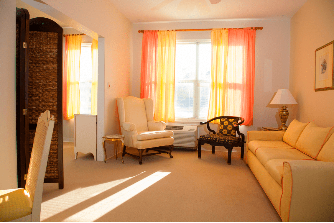 A cozy living room with warm sunlight streaming through orange curtains, featuring an armchair and a small side table.
