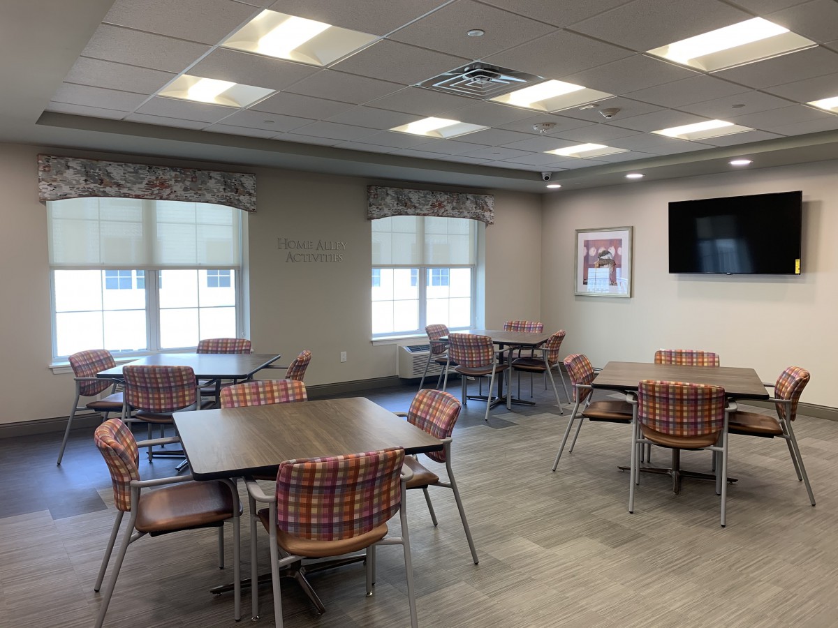 An empty cafeteria with several tables and chairs, featuring windows and a television on the wall.