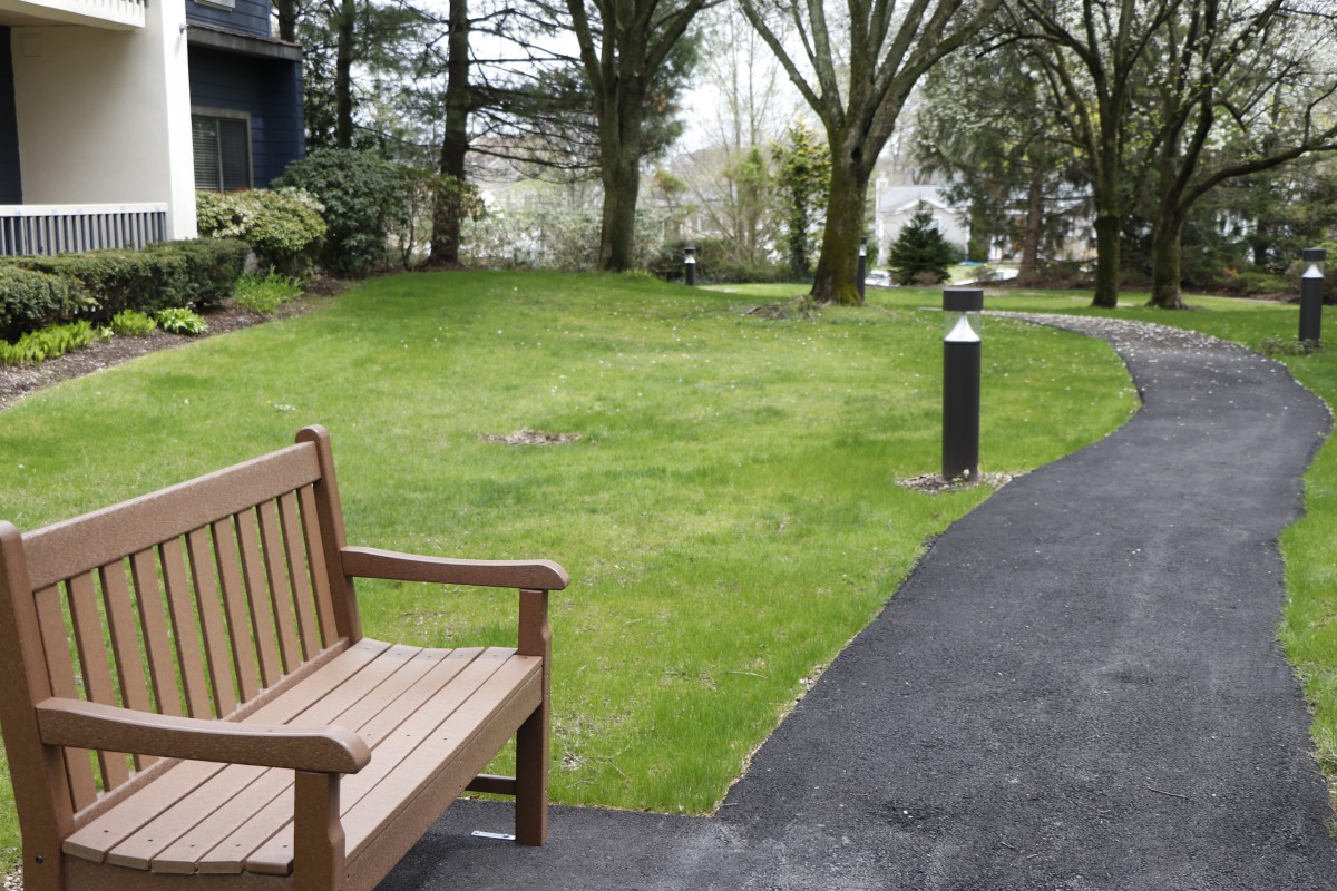 A tranquil outdoor garden area with a wooden bench and a paved pathway surrounded by green grass and trees.