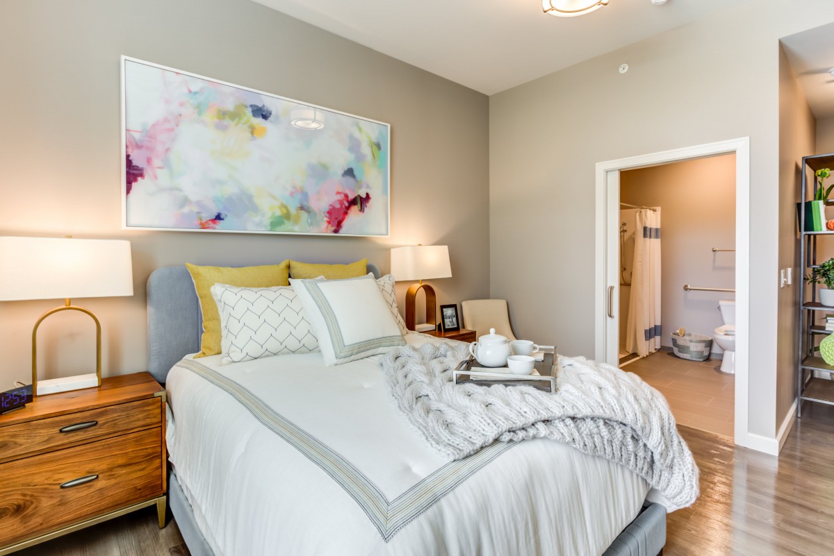 A cozy and modern bedroom with elegant decor, featuring a neatly made bed, colorful artwork above the headboard, and warm lighting from stylish lamps.