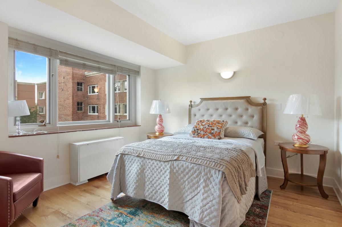 A cozy bedroom with a neatly made bed, adorned with decorative pillows. Natural light flows through the window, illuminating the soft-colored walls and wooden flooring. A lamp sits on the nightstand beside the bed.