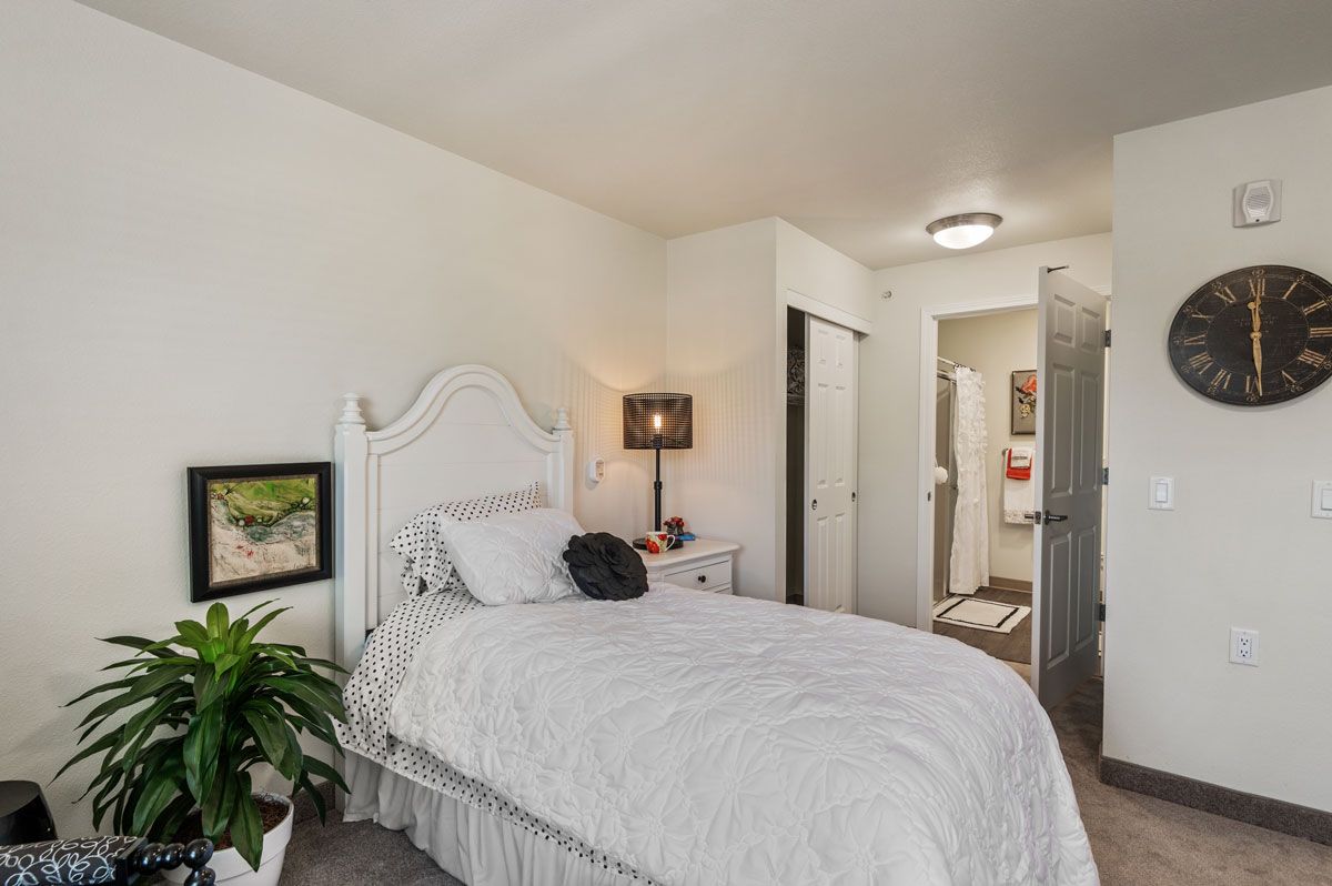 A cozy bedroom with white walls, featuring a neatly made bed, a small bedside table with a lamp, and decorative elements like a plant and framed artwork.