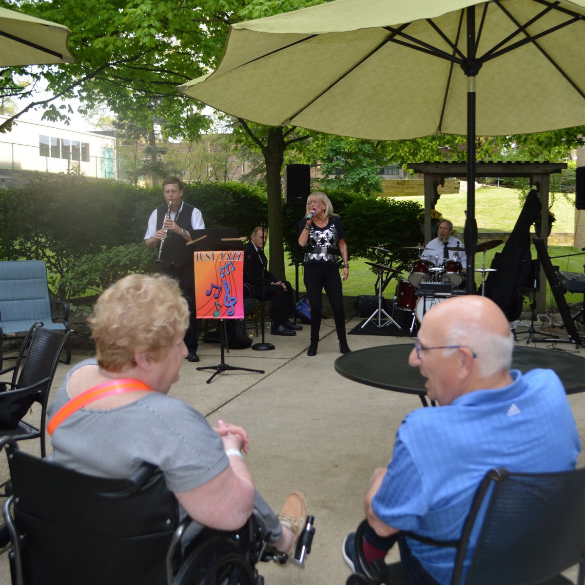 An outdoor event with musicians performing under umbrellas, while an audience watches from their seats.