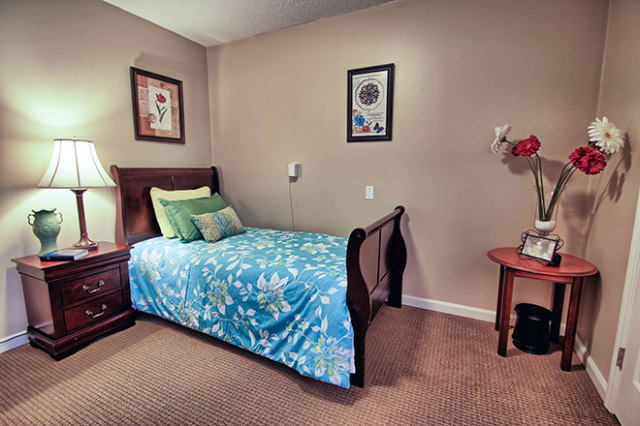 A cozy bedroom with a single bed featuring blue floral bedding, adorned with decorative pillows. The room is complemented by framed artwork on the walls and a wooden nightstand with a lamp.