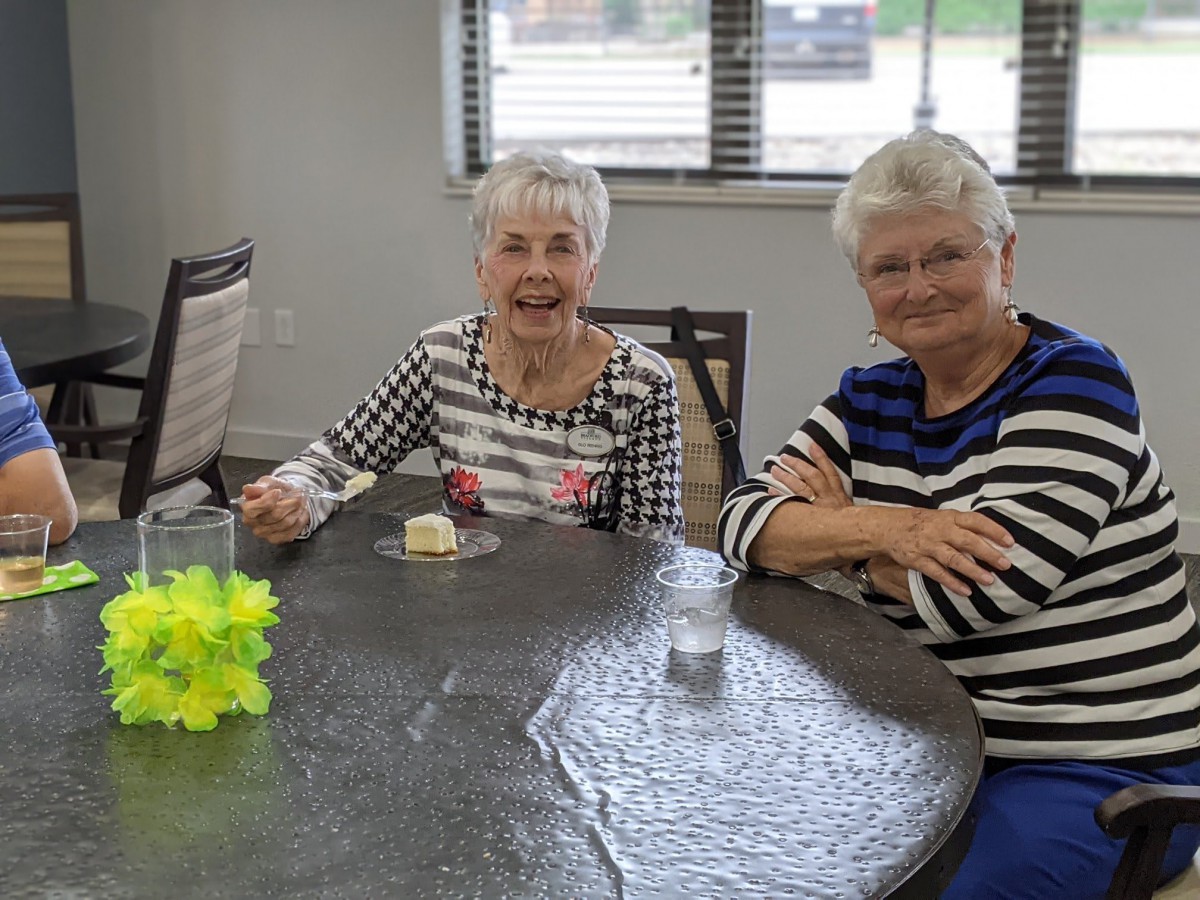 Two elderly women smiling and seated at a table indoors.