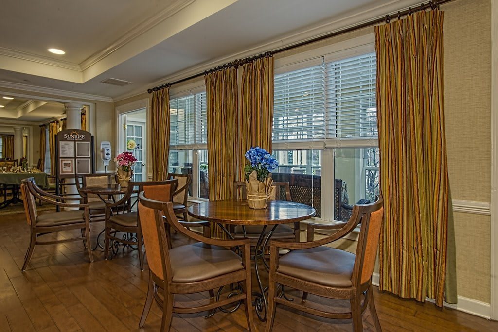 A cozy dining area with wooden tables, chairs, and large windows adorned with curtains.