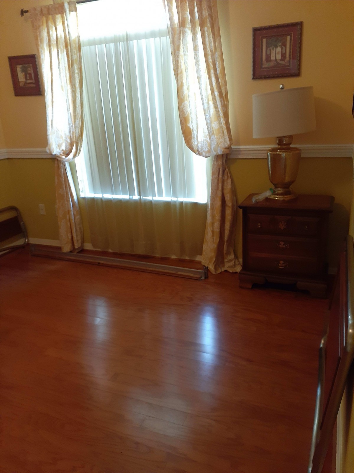 An empty room with hardwood floors, a window with curtains, and a dresser with a lamp on top.