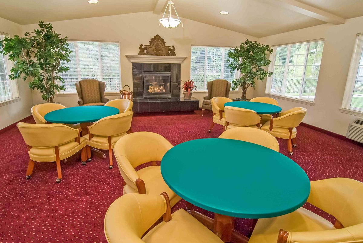 A spacious lounge area featuring multiple round tables and chairs, centered around a fireplace with large windows and indoor plants.
