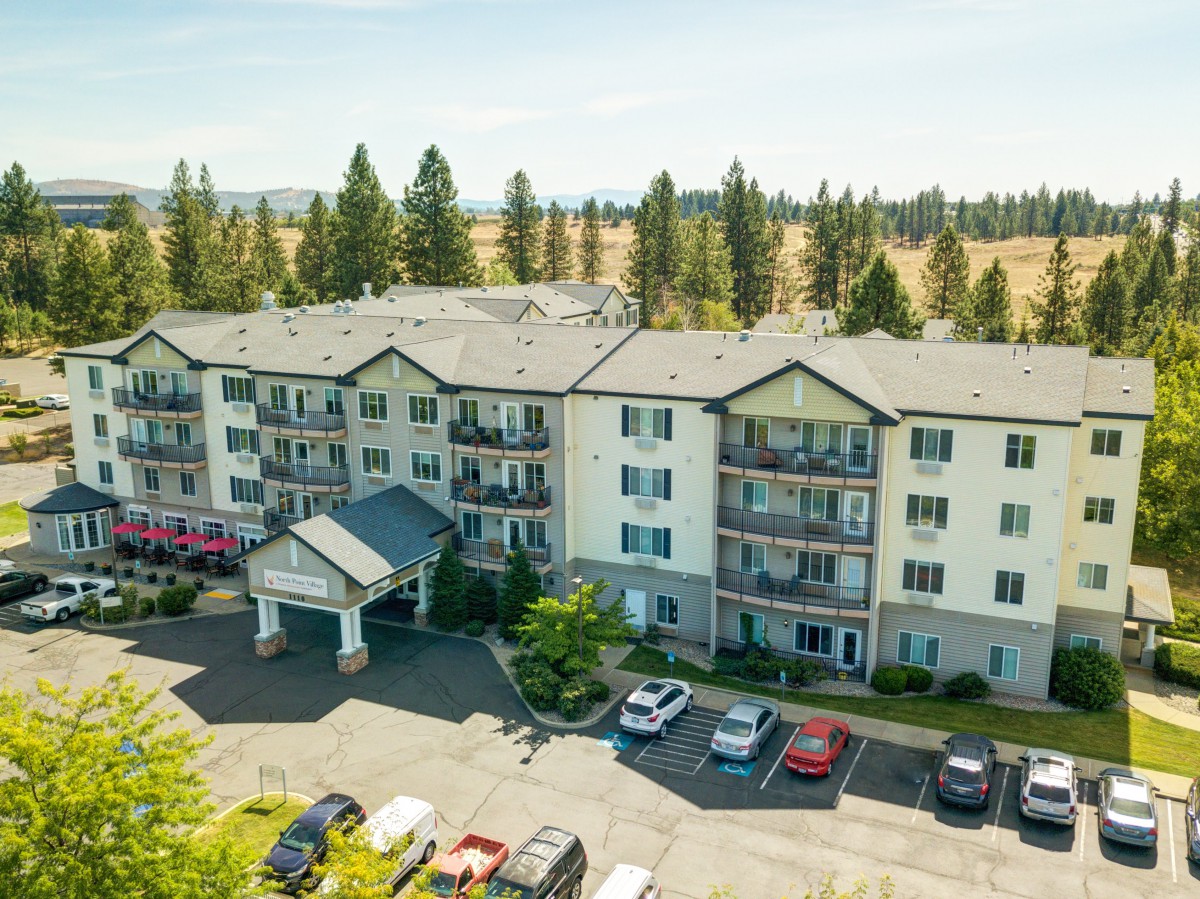 Aerial view of a residential apartment building with a parking lot in front, surrounded by trees and greenery.