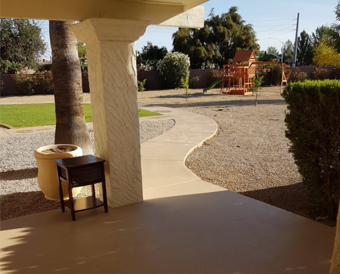 A sunny backyard viewed from a patio, featuring a pathway and garden furniture.