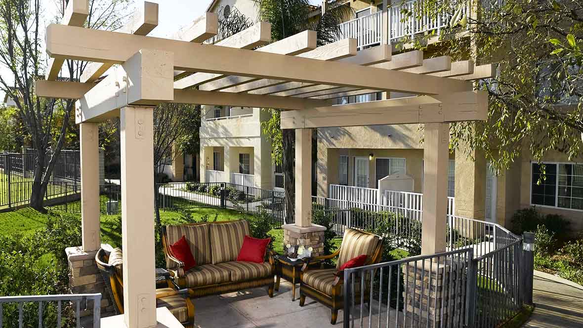 A cozy outdoor patio with a pergola, featuring comfortable seating and surrounded by apartment buildings and greenery.