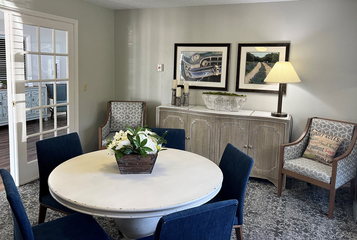 A cozy living room with a round table surrounded by blue chairs, decorated with artwork and flowers.