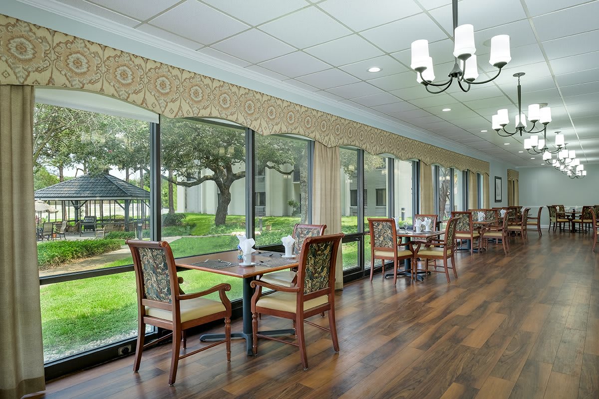 A well-lit dining area with large windows, featuring wooden floors and a view of the greenery outside.