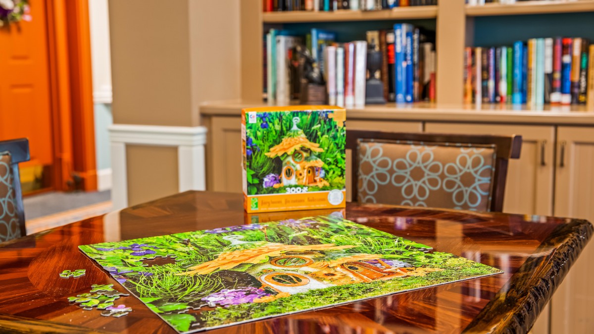 A completed jigsaw puzzle featuring a house in the woods sits on a table, with its box and a bookshelf in the background.