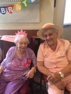 Two elderly women smiling, wearing hats and enjoying a celebration indoors.
