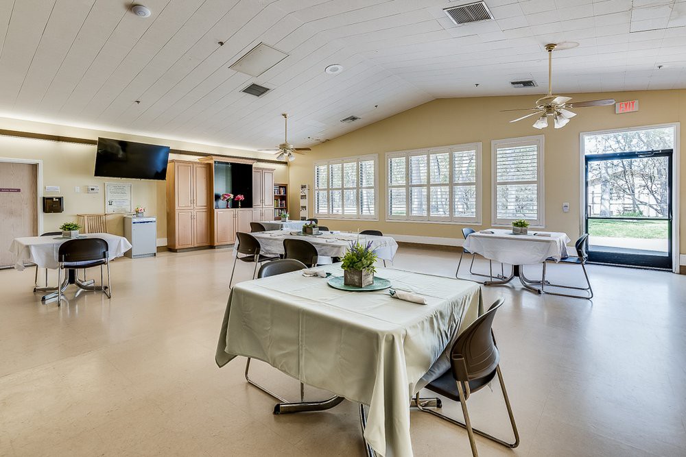 A spacious dining area with neatly arranged tables and chairs, featuring large windows and ceiling fans.