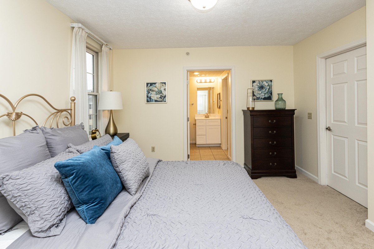 A cozy bedroom with a neatly made bed, decorative pillows, and a view into an adjoining bathroom.