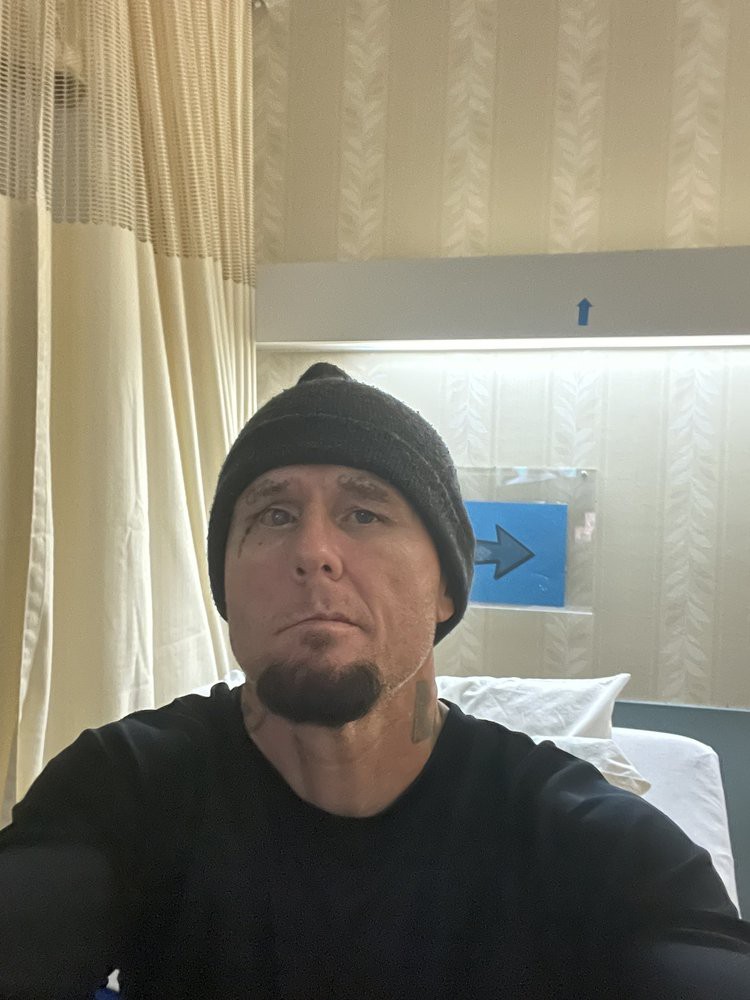 A man wearing a black beanie is taking a selfie in a room with curtains and a bed.