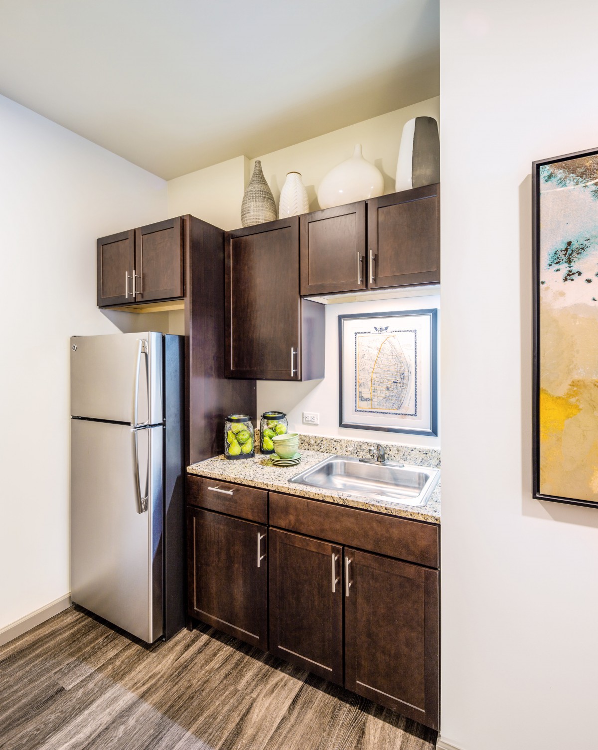A modern kitchen with dark wood cabinets, a stainless steel refrigerator, and decorative artwork on the wall.