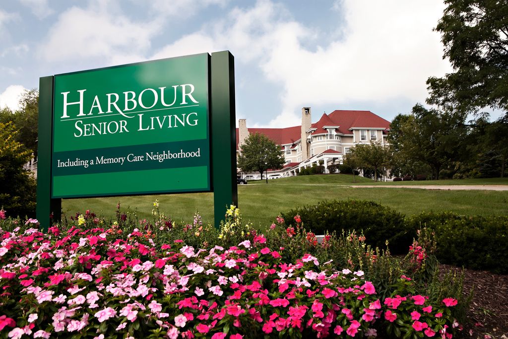A scenic view of a senior living facility with colorful flowers and a welcoming sign.