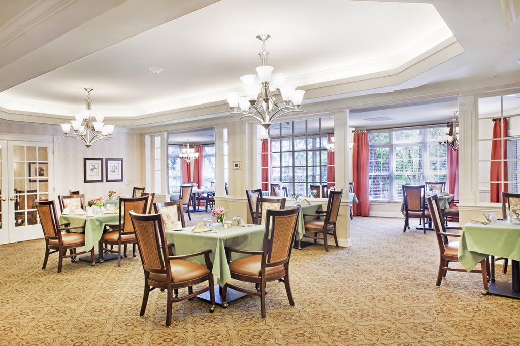 A spacious and elegantly decorated dining room with multiple tables, chairs, and chandeliers, illuminated by natural light from large windows.