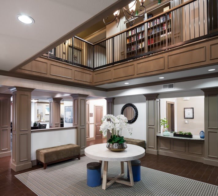 A modern lobby with a central round table, decorative plants, and an upstairs bookshelf area.